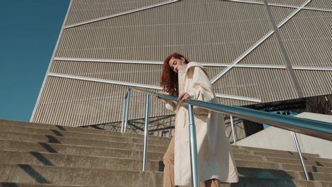Young Red-Haired Girl On The Street. Girl Has Long Curly Hair. Urban Fashion Concept. Girl Walks Up The Stairs Near Modern Building.