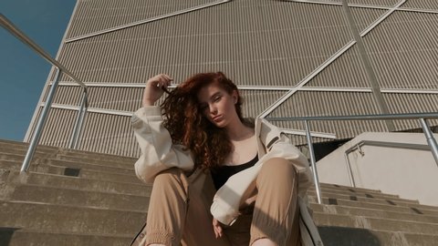 Young Red-Haired Girl On The Street. Girl Has Long Curly Hair. Urban Fashion Concept. Pretty Girl In Raincoat And Sneakers Sits On The Stairs Of Modern Building. Girl Playing With Her Hair.