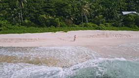 Aerial footage of young tanned woman running on sandy tropical beach with ocean waves rolling. Mahe island. Seychelles. Travel lifestyle concept