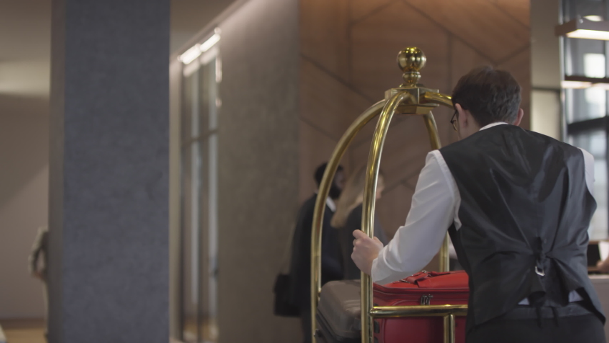 Tracking with rear view of male hotel bellhop in uniform wheeling cart full of guests luggage Royalty-Free Stock Footage #1045879033
