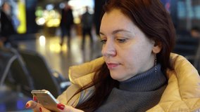 A 30-35-year-old woman has chat correspondence with someone sitting in a chair while waiting for boarding a flight at the airport. The concept of tourism and flight delay.