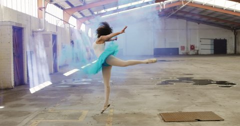Side view of mixed race female dancer practicing in an empty warehouse, dancing, turning around and holding smoke grenade, slow motion