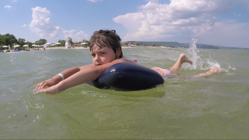 Wave cover the child. A little kid swims on the inflatable mattress. Child shows her tongue.