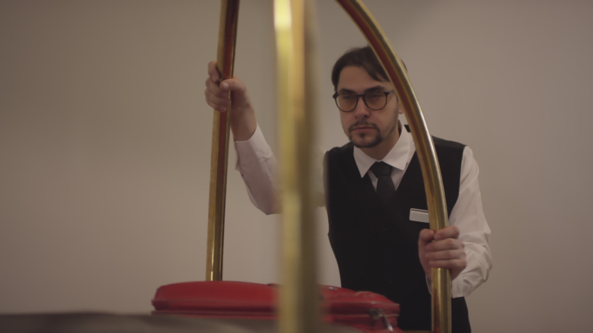 Tracking shot of focused bellman in glasses and uniform pushing luggage cart along hallway and through lobby of hotel Royalty-Free Stock Footage #1045888177