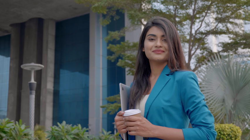 An Indian woman or lady executive with a laptop and a coffee mug in hand laughing facing the camera. A happy corporate female employee smiling looking at the camera in front of a corporate building. Royalty-Free Stock Footage #1045888723