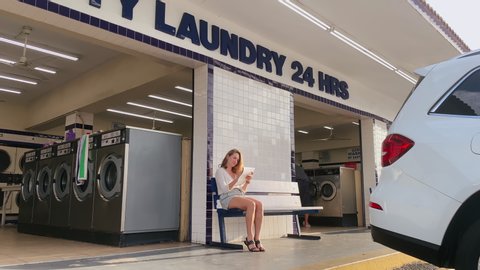 Young attractive woman waiting for laundry outside near entry to launderette in summer. Housewife spending time in public laundry