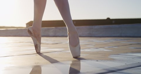 Close up of legs of a female ballet dancer practicing on rooftop at sunrise, walking on tiptoes and jumping, buildings in the background, slow motion