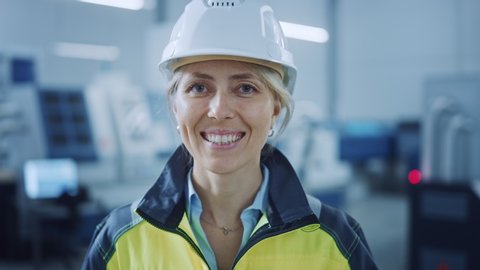 Portrait of Beautiful Smiling Female Engineer Wearing Safety Vest and Hardhat Takes of Safety Goggles. Professional Woman Working in the Modern Manufacturing Factory. Facility with CNC Machinery