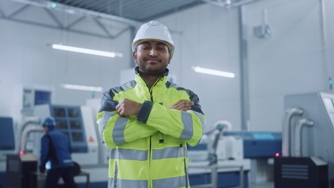 Portrait of Handsome Indian Engineer Wearing Safety Vest and Hardhat Crosses Arms and Smiles. Professional Woman Working in the Modern Manufacturing Factory. Facility with CNC Machinery and robot arm