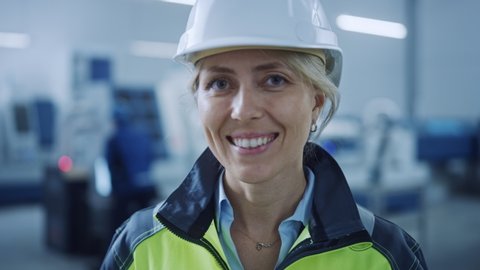 Portrait of Beautiful Smiling Female Engineer Wearing Safety Vest and Hardhat Takes of Safety Goggles. Professional Woman Working in the Modern Manufacturing Factory.  CNC Machinery and Robot Arm