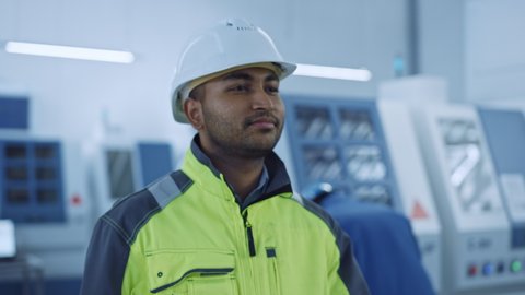 Industry 4 Factory: Portrait of a Modern Worker Wearing Safety Jacket Puts on Hard Hat and Walks Through Contemporary Industrial Workshop where Professionals are Working and Programming CNC Machinery