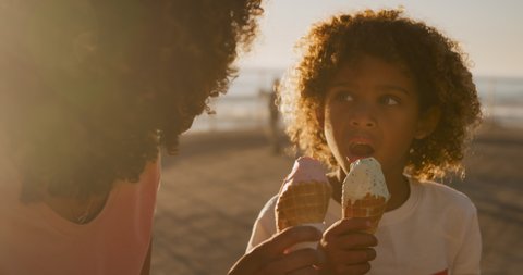 Front view close up of mixed race woman with her son enjoying family time by the seaside together on a sunny day, eating ice cream, slow motion 