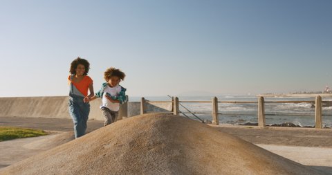 Front view of mixed race woman with her son enjoying family time by the seaside together on a sunny day with blue sky, running, holding hands, slow motion 庫存影片