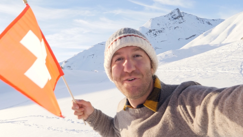 Selfie of young man holding Swiss flag surrounded by mountain peaks in the Alps 