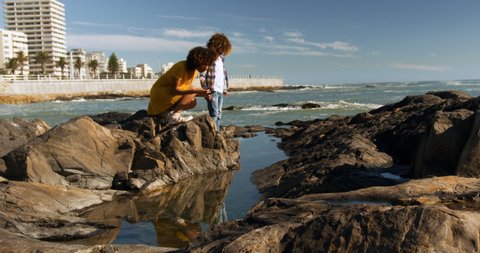 Side view of mixed race woman with her son enjoying family time by the seaside together on a sunny day with blue sky, kneeling down, looking at a rock pool and pointing, slow motion