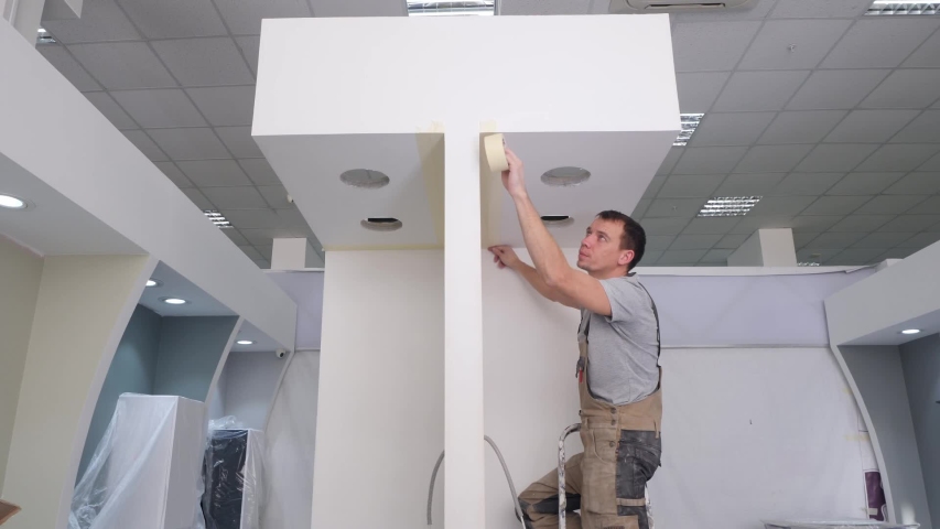 professional handyman in jumpsuit glues masking tape on new apartment wall standing on metal ladder
