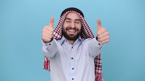 Bearded young arabian muslim man 20s in keffiyeh kafiya ring igal agal casual clothes posing isolated on pastel blue background. People religious lifestyle concept. Looking at camera, showing thumb up