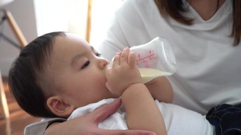 Close-up of Asian mother babysitting and feeding her own son with milk bottle in living room at home. - Motherhood and formula-fed newborn concept