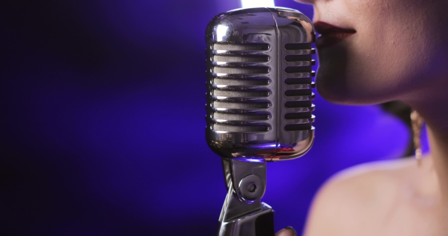 Singer is performing a song on concert. Woman is singing in microphone on blue background, closeup lips side view. Performance on musical concert on event or party.
