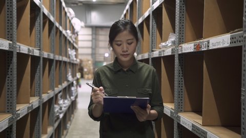 Candid of young attractive asian korean woman auditor staff work walking looking up stock taking inventory in warehouse by writing on clipboard making notes. female owner and small business concept.
