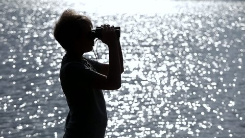 Black silhouette of young curious white kid holding binoculars in hands and looking at distance. Boy isolated at blurry sparkling sunny blue river water background.