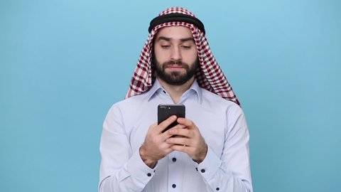 Bearded young arabian muslim man 20s in keffiyeh kafiya ring agal casual clothes isolated on pastel blue background. People religious lifestyle concept. Looking surprised wow, using mobile cell phone