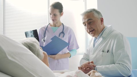 Caring doctor talking with a senior patient and supporting her, a nurse is smiling and standing next to them