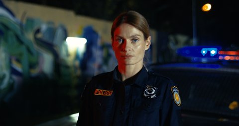 Portrait of attractive Caucasian young female police officer looking in car at night. Policewoman at the police car withh lights outdoor in dark. Quarantine coronavirus infection, protection