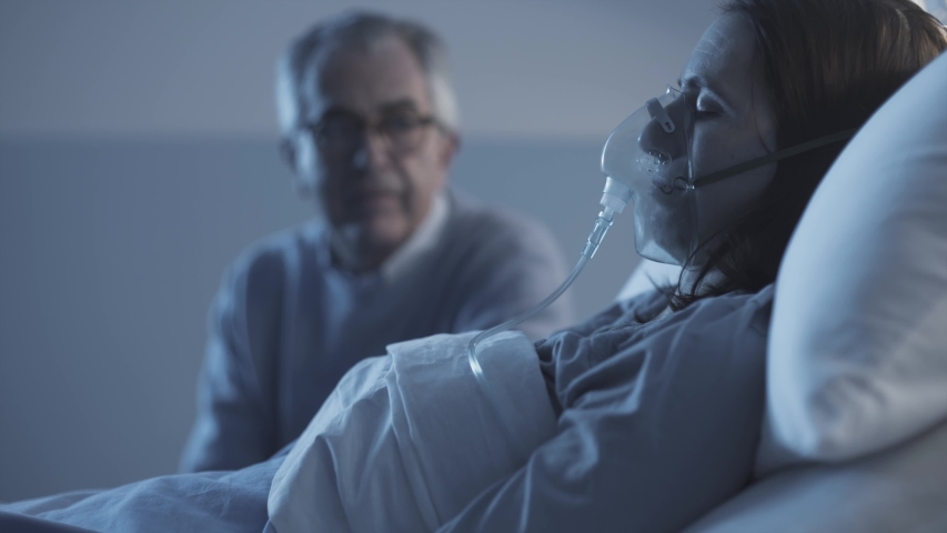 Caring man checking a sick woman sleeping in a hospital bed with an oxygen mask