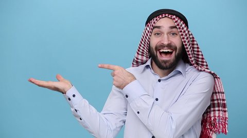 Bearded young arabian muslim man 20s in keffiyeh kafiya ring igal agal casual clothes posing isolated on pastel blue background. People religious lifestyle concept Pointing fingers on copy space place