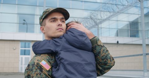 Portrait of military man holding son in his arms and stroking hair. Young soldier embracing his boy kid before living on military duties.Concept of family connection military service. Outdoors