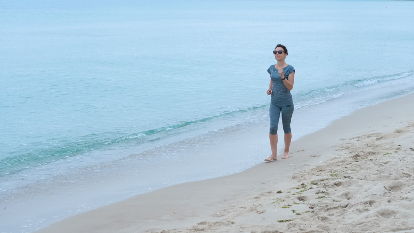 Fitness female near the sea. A sporty women with bare foot runs on the sandy beach, female stops, looks at her watch and continues to flee further.