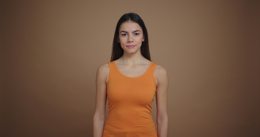 Young beautiful woman standing isolated on brown background, smiling modestly and looking on camera