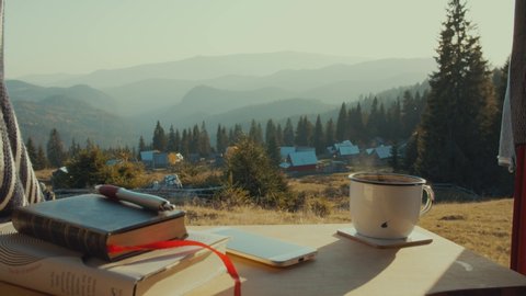 Van Life shot of steaming coffee plus notebook and cellphone in front of mountain range panorama at dusk on a hill with forests in Romania
