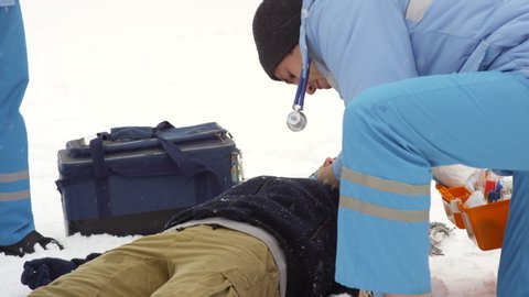 Sequence of shots of team of paramedics arriving at scene of accident and running towards eyewitnesses gathered around unconscious patient lying on snow, then checking their vitals