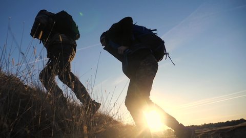 two tourists teamwork with backpacks slow motion walking go silhouette in the sunlight glare of the sun at sunset. two men hikers climb lifestyle mountains overcoming difficulties concept teamwork