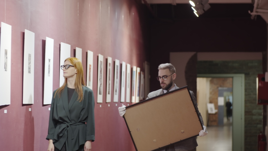 Male gallery worker in white cotton gloves showing framed painting to female manager and discussing it with her while working in exhibition hall