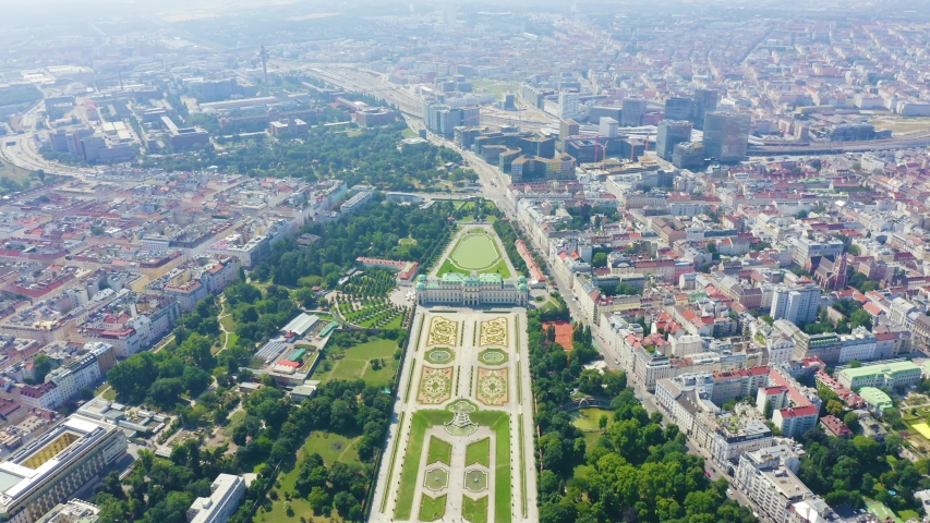 Vienna, Austria. An aerial view captures the sprawling urban landscape dominated by the Belvedere, a baroque palace complex consisting of the Upper and Lower Belvedere set within extensive geometrically patterned gardens. The grand palaces feature ornate facades and are surrounded by dense green parkland. Beyond the palace grounds, the city extends with a mix of low-rise residential buildings featuring red-tiled roofs and modern office towers. A major highway and railway lines cut through the city in the background. The scene is bathed in bright summer daylight with a clear blue sky, highlighting the contrast between historic architecture and contemporary urban development. Departure of the camera. Aerial view