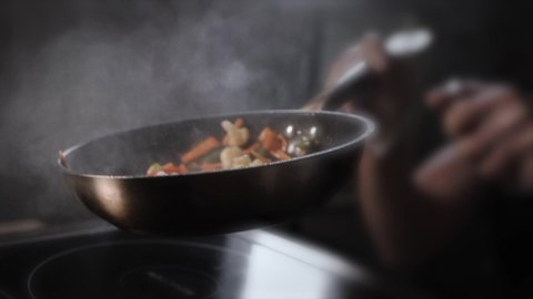 Cook in transparent sterile gloves in the kitchen throws up a hot frying wok with chicken, vegetables and noodles in a black pan