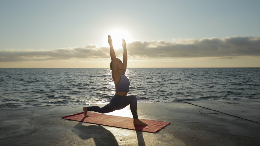 A young girl does a yoga exercise, moving from one pose to another, on a pier in the morning sun on a background of the sea.