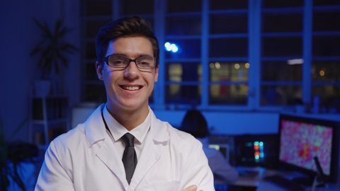 Tracking medium shot of handsome young chemist in eyeglasses and white lab coat standing in laboratory looking at camera and smiling happily, his female colleague working on laptop in background