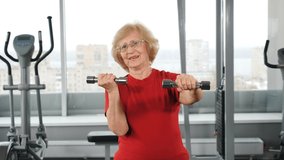 Active senior retired woman with barbells in gym. Active and happy old years concept. Positive retired person making light exercises with dumbbells in fitness club. 4k video