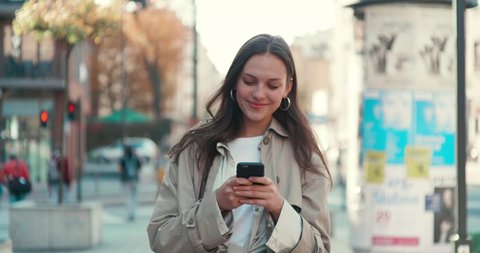 Young Attractive Girl with Long Brunette Hair walking in the Street, Having Mobile Phone in her Hands. Typing Messages and Smiling while sending. Having fashionable clothes and Stylish earrings.