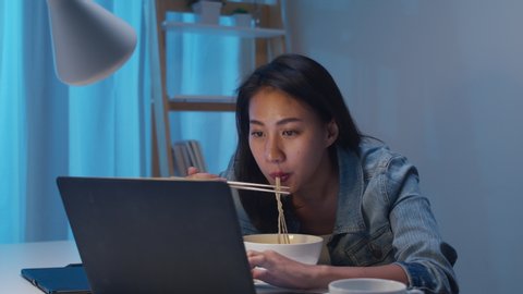 Asia freelance smart business women eating instant noodles while working on laptop in living room at home at night. Happy young Asian girl sitting on desk work overtime, enjoy relax time.