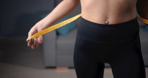 Close up of young woman with slender body measuring waist with tape after morning workout at home. Fitness lady with bare torso estimating new forms of her figure.