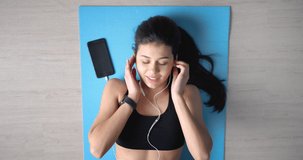 Top view of cheerful woman with dark hair lying on yoga mat, listening music on earphones and singing along. Attractive lady with perfect body shape in sport clothing having fun during workout.
