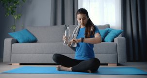 Attractive woman with slim body sitting on blue yoga mat with crossed legs, drinking water and looking at her smart watch. Athletic girl in sport clothing resting between exercises at living room.
