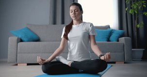 Attractive sports women with dark hair sitting on yoga mat with closed eyes in pose lotus at living room. Young fitness girl meditating during morning workout.