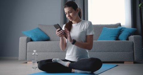 Attractive young lady in sports wear sitting on yoga mat with crossed legs and using smartphone after trainings. Relaxing woman having break at living room with grey and blue interior colors.
