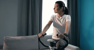 Motivated young girl in sportswear doing exercise on bicycle at home and listening music on earphones. Charming brunette training everyday to stay fit and healthy